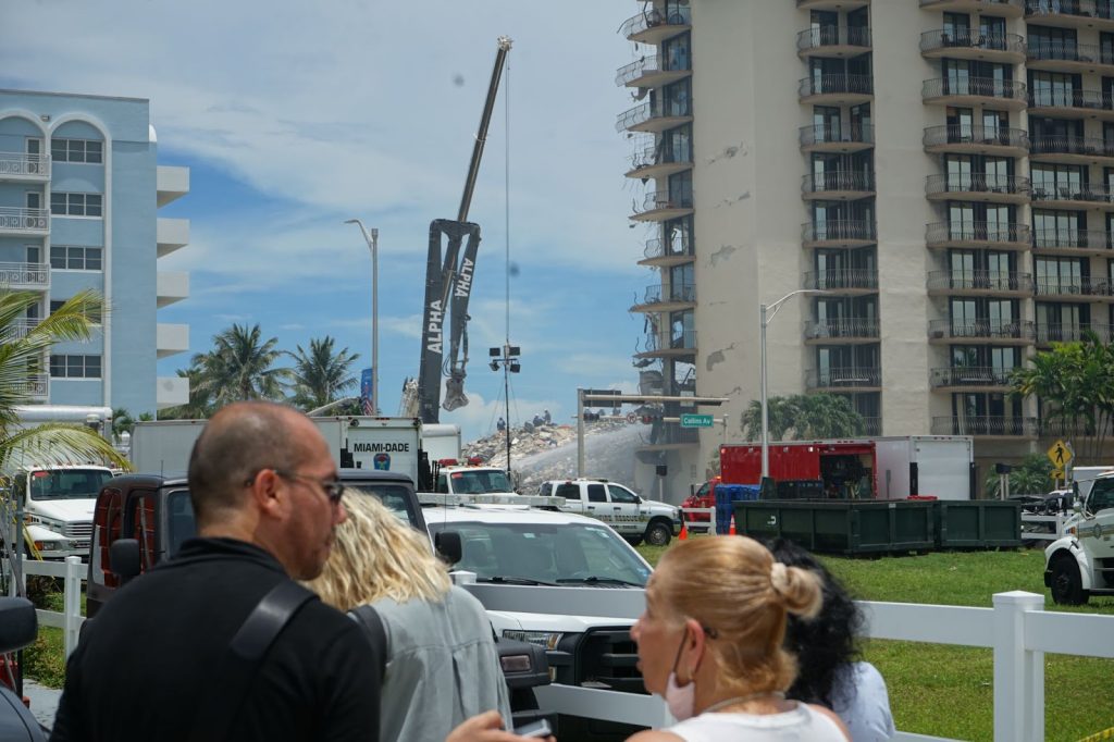 Spectators observe Saturday’s continued rescue mission as first responders slowly work through debris on Saturday, June 27. The surviving section of Champlain Towers South stands to the right, coated in chipping paint and dust.
