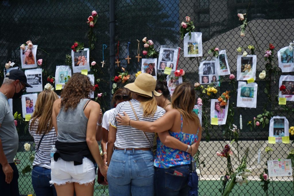 A family huddles close at the Surfside Tennis Center on Saturday, June 26. The center's gate has been used as a memorial wall for those lost in the collapse.
