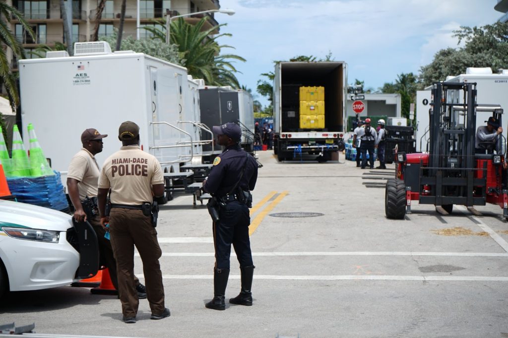 Miami Dade police officers make conversation outside of an improvised restroom area at the site of the collapse. Volunteers and first responders work and talk in the background on Saturday, June 26, the third day of rescue operations.